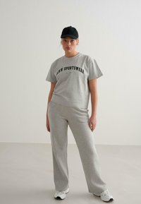 Šedé bavlněné tričko s nápisem "ADMN' SPORTSWEAR", doplněné o šedé tepláky a černou čepici. Sportovní bílé tenisky dotvářejí vzhled.