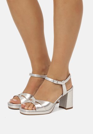 Pies calzados con sandalias plateadas de textura, tacón bloque, con correas y hebillas en el tobillo, mostrados contra un fondo claro y liso.