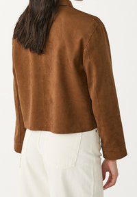 Bruine suède cropped jas met een gladde textuur, voorzien van een kraag en lange mouwen. Het model draagt een witte broek en toont het achteraanzicht.