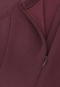 Bordeauxfarvet lynlås i stof med en glat struktur og synlige syningsdetaljer. Lynlåsen er placeret langs kanten af tøjet.