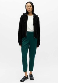 Black textured zip-up cardigan over a white top, paired with green tapered pants and black loafers. Simple and modern design.
