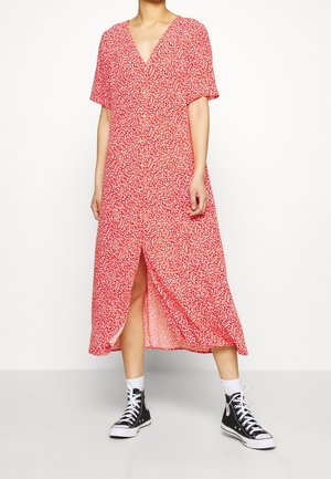 Vrouw draagt een rode midi-jurk met knopen en een witte bloemenprint, witte sokken en zwarte hoge sneakers, staande op een effen achtergrond.