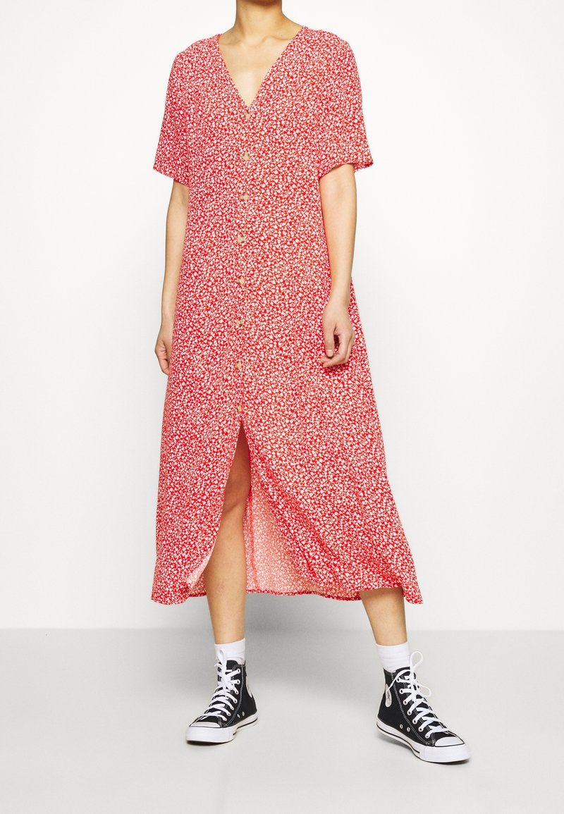 Femme portant une robe midi rouge à boutons avec un imprimé floral blanc, des chaussettes blanches et des baskets montantes noires, debout sur un fond uni.