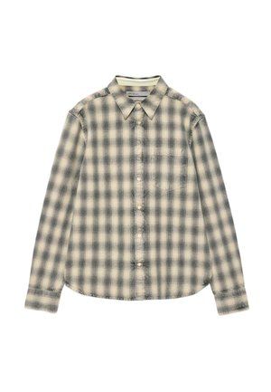 Joven de pie con una camisa de manga larga beige y negra a cuadros, vaqueros azules y zapatos de ante marrones sobre un fondo liso.