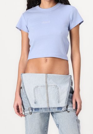 Kvinde iført en lyse lilla cropped t-shirt og lyseblå denimbukser med et aftageligt denimpanel ved taljen.