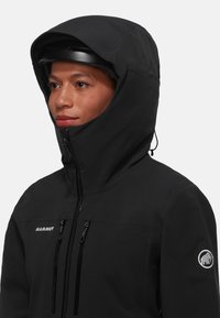 Svart Mammut-jacka med hög krage och fast huva. Har två fickor med dragkedja på bröstet och en logotyp-patch på ärmen.