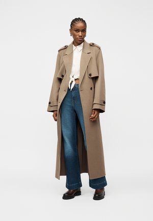 Femme aux cheveux tressés portant un long trench-coat beige, une chemise blanche nouée, un jean bleu à jambes larges, et des bottes marron foncé, debout devant un fond uni.