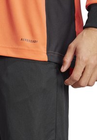Aeroready Performance-Shirt mit orange und schwarzen Einsätzen, glatter Textur, flachen Nähten und Logo in der Nähe des Saums. Schwarze Hose mit sichtbarem Taschen.