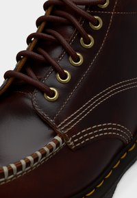 Κοντινή λήψη σκούρου καφέ δερμάτινης μπότας με κίτρινες οπές, καφέ κορδόνια, λεπτομερή μπεζ ραφή και μαύρη σόλα με κίτρινες λεπτομέρειες.