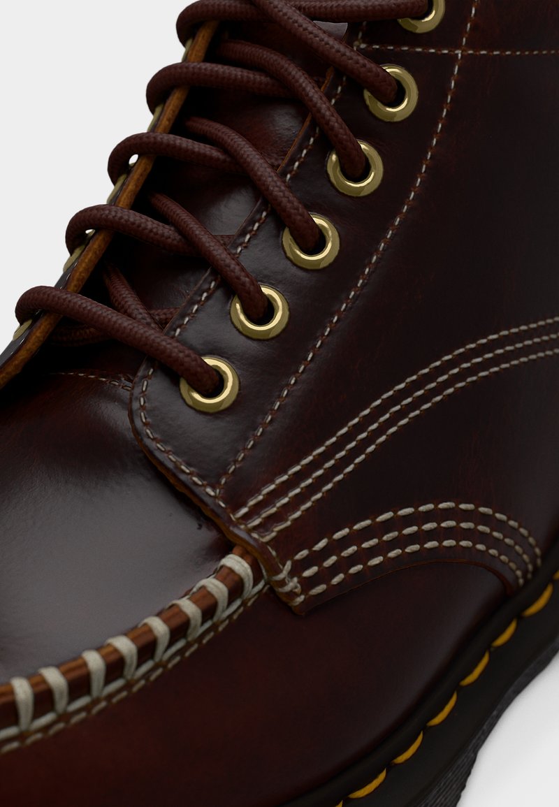 Close-up van een donkerbruine leren laars met gele veteroogjes, bruine veters, gedetailleerd beige stikwerk en een zwarte zool met gele accenten.