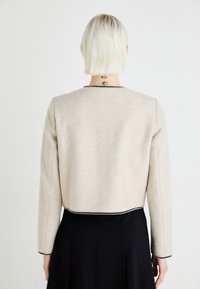 Beige cropp jacket met lange mouwen, voorzien van een zwarte geborduurde rand langs de halslijn en de zoom. Zachte, gestructureerde stof.