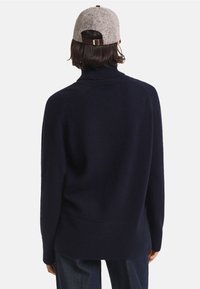 Pulover din lână de culoare bleumarin, cu gât înalt și croială lejeră, cu manșete și tiv cu țesătură în relief, asortat cu o șapcă gri deschis cu o curea maro.