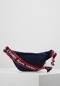 Marsupio in tessuto blu navy con cinturino rosso e bianco "TOMMY JEANS", dotato di chiusura con zip e dettagli in metallo argentato.