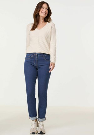 Cremefarbener V-Ausschnitt-Pullover mit diagonaler Rippstruktur, kombiniert mit dunkelblauen Jeans, die an den Säumen aufgerollt sind, und beigen Sneakers.