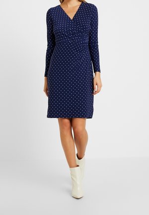 Vrouw draagt een marineblauwe lange mouwen jurk met witte stippen en crèmekleurige enkellaarzen, staand tegen een effen lichte achtergrond.