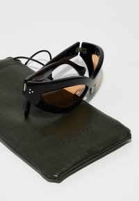 Occhiali da sole neri con forma a occhio di gatto, lenti color marrone e finitura lucida. Accompagnati da una custodia in pelle verde scuro con texture.