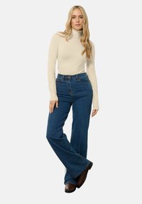Beige Rollkragenpullover kombiniert mit hoch taillierten, ausgestellten blauen Jeans und braunen Stiefeln. Der Stoff wirkt weich mit einer glatten Oberfläche.