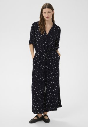 Donna con lunghi capelli castani che indossa una tuta blu navy con pois, maniche corte e scarpe basse nere, con le mani in tasca.