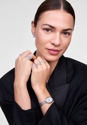 Montre en maille argentée avec un cadran rond, associée à plusieurs bagues argentées et des boucles d'oreilles créoles, présentée sur un blazer noir.