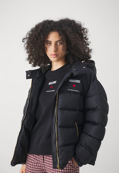 Crna puffer jakna s visokim ovratnikom i zlatnim patentnim zatvaračima. Sadrži izvezeni Moschino logo i akcentne motive srca. Nošena preko crnog duksera.