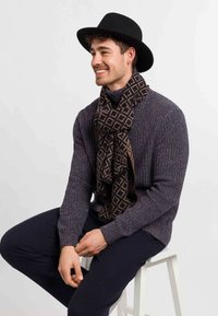 Un homme souriant portant un chapeau noir à large bord, une écharpe à motif, un pull en laine violet et un pantalon sombre est assis sur un tabouret blanc devant un fond uni.