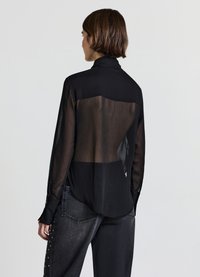 Schwarze durchsichtige Bluse mit hohem Kragen und langen Ärmeln, die einen halbtransparenten Rücken und einen glatten, strukturierten Stoff aufweist. Kombiniert mit dunklen Jeans.