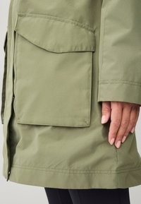 Olijfgroene jas met een gladde textuur, voorzien van een grote voorkantzak en een rechte zoom, getoond met een hand die erop rust.