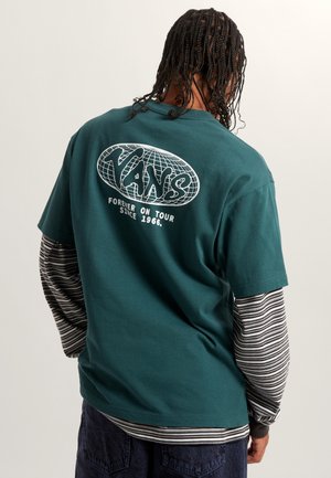 Tee in Türkis aus Baumwolle mit einem weißen Globus-Graphic, darunter der Text "VANS" und "Forever on Tour since 1966", über einem gestreiften Langarmshirt getragen.