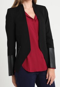 Svart kavaj med figursydd design, som har dragkedjefickor och läderdetaljer på ärmarna. Bärs över en vinröd blus.