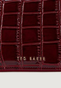 Burgunderfärgad lackläderhandväska med en quiltad textur och rutmönster. Guld "TED BAKER"-logotyp präglad på framsidan.