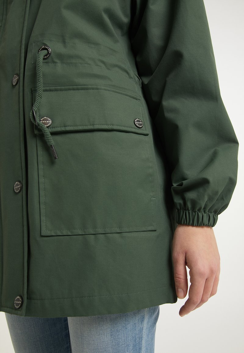 Grüne wasserdichte Jacke mit einem Reißverschluss vorne und Druckknöpfen, ausgestattet mit einem verstellbaren Kordelzug in der Taille und elastischen Bündchen. Zwei Fronttaschen.