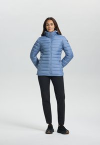Femme se tenant avec les mains dans les poches, portant une veste matelassée bleu clair, un pantalon noir et des baskets noires sur un fond blanc.