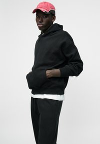 Musternööbiga must kapuutsiga sviiter, millel on ees tasku, kantud valge särk üle. Kombineeritud mustade dressipükstega ja roosa kulunud kübaraga. Lihtne ja mugav disain.