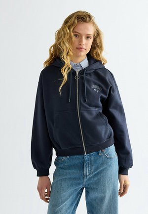 Giovane donna con capelli biondi mossi che indossa una felpa con cappuccio blu navy con zip sopra una maglietta a righe e jeans azzurri su uno sfondo chiaro uniforme.