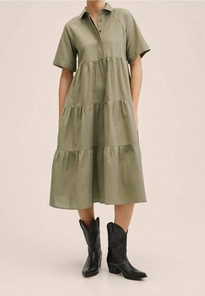 Mujer con un vestido de volantes hasta la rodilla de color verde oliva con mangas cortas y botas negras de estilo vaquero hasta la mitad de la pantorrilla, de pie frente a un fondo liso.