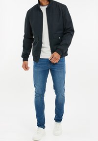 Man med svart zip-up jacka, ljusgrå skjorta, blå jeans och vita sneakers som står mot en vit bakgrund.