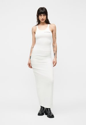 Femme mince aux longs cheveux noirs et tatouages porte une robe maxi blanche sans manches ajustée et des bottes plateformes noires sur un fond blanc.