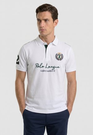 Hombre con cabello castaño que lleva una camiseta blanca de Polo League con logotipos verdes y pantalones azul marino, de pie con las manos en los bolsillos, mirando hacia un lado.
