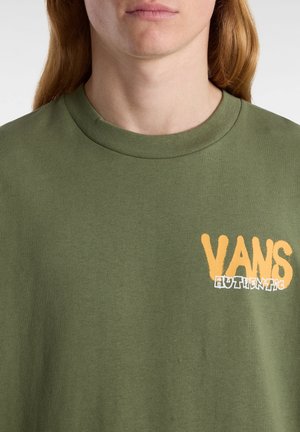 Nahaufnahme einer Person, die ein olivgrünes T-Shirt mit Rundhalsausschnitt trägt, auf dessen Brust sich das orange-weiße "VANS Authentic"-Logo befindet.