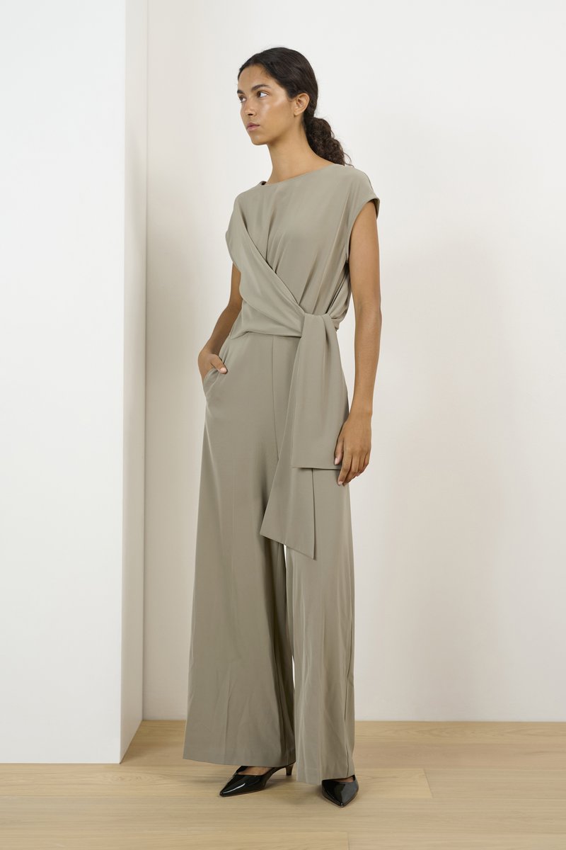 Khaki jumpsuit with a wrap design, short sleeves, side pockets, and a tied waist detail. Textured fabric and wide-leg silhouette. Black heels.