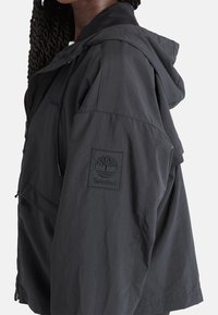 Giacca leggera nera con cappuccio, caratterizzata da una toppa con logo texturizzato sulla manica, tasche con cerniera e un design a vestibilità rilassata.