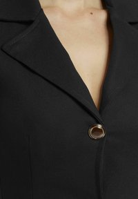 Schwarzer Blazer mit Reverskragen, einem einzelnen Knopfverschluss mit goldener Akzentuierung und einer strukturierten Stoffoberfläche.