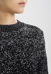Schwarz-weiß gemusterter Strickpullover mit geripptem Halsausschnitt, einer strukturierten Oberfläche und legerer Passform, von der Schulter aufwärts gezeigt.