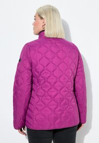 Žena s krátkými blond vlasy, oblečená v prošívané fialové bundě s geometrickým stehem, stojící na pozadí čistě bílé.