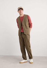 Olivgrüner Weste über einem roten strukturierten Pullover, kombiniert mit olivfarbenen Hosen und weißen Sneakers mit Gummisohle. Das Model trägt eine beigen Mütze.
