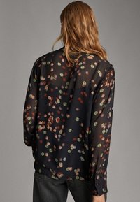 Kvinna med långt ljusbrunt hår som bär en genomskinlig svart blus med ett mönster av flertalet färgglada cirkelfläckar och mörka byxor, vänd bort.