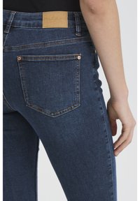 Tuvplāns personas, kas valkā tumši zilas šauras džinsas ar aizmugurējo kabatu un brūnas ādas "PULZ JEANS" etiķeti uz jostasvietas.