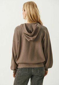 Brown oversized hoodie in soft fabric with a hood, ribbed cuffs, and hem. Paired with dark gray jeans, viewed from the back.