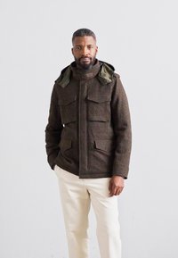 Veste en cuir marron texturé avec une fermeture éclair devant, quatre grandes poches et une capuche amovible vert olive, accompagnée de pantalons de couleur crème.