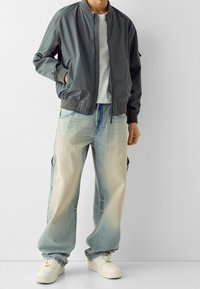 Grå bombarjacka med ribbade ärmar, tillsammans med ljusa, urtvättade baggyjeans och vita sneakers. Outfiten har en avslappnad och ledig passform.